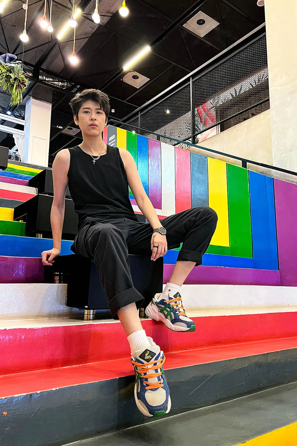 Lesbian-presenting fashionista rocks TOMSCOUT’s compression tank and black cuffed trousers, lounging effortlessly on colorful stairs framed by Progress Pride hues—a radiant tribute to queer joy and self-expression.