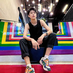 Tomboy model sits on rainbow-striped stairs beneath hanging lights, dressed in TOMSCOUT’s sleek black binder and cuffed pants, embodying bold queer confidence with every unapologetic stare.