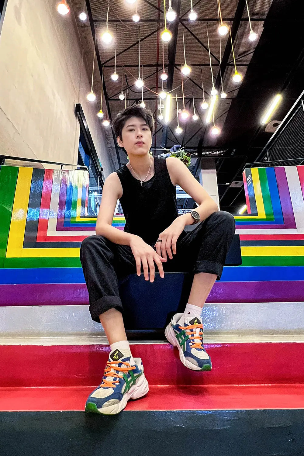 Tomboy model sits on rainbow-striped stairs beneath hanging lights, dressed in TOMSCOUT’s sleek black binder and cuffed pants, embodying bold queer confidence with every unapologetic stare.