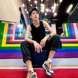 Tomboy model sits on rainbow-striped stairs beneath hanging lights, dressed in TOMSCOUT’s sleek black binder and cuffed pants, embodying bold queer confidence with every unapologetic stare.