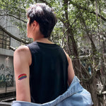 Close-up of a non-binary person with a short fade haircut, wearing a sleeveless black tee and denim jacket draped off the shoulders, revealing a rainbow Pride tattoo on their arm—an unapologetic symbol of LGBTQ+ identity and personal liberation.