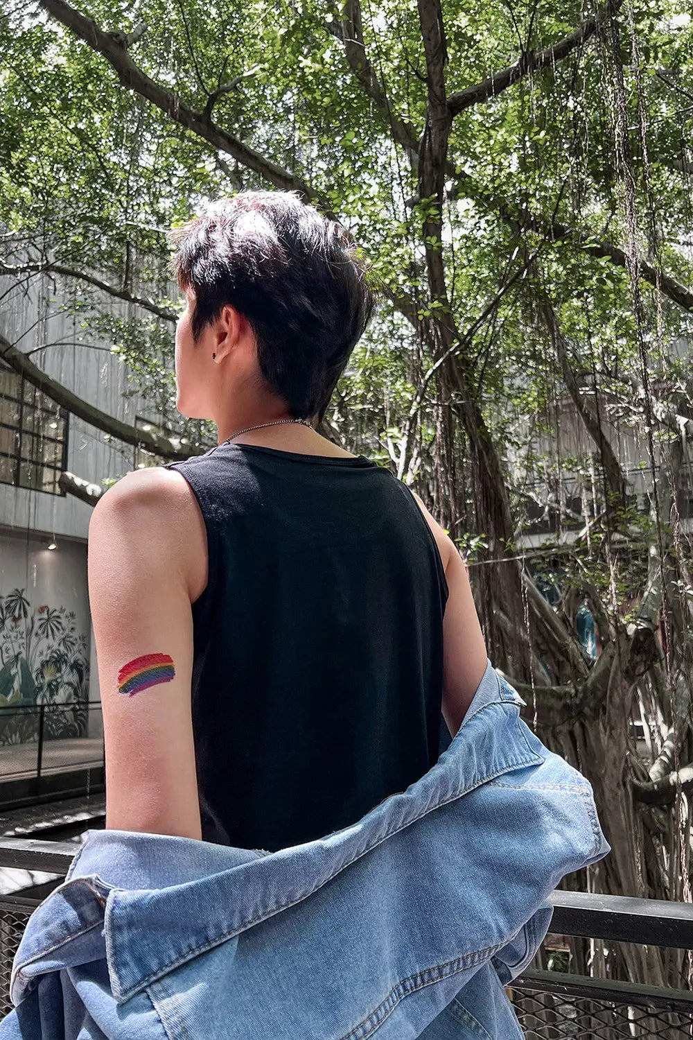 Close-up of a non-binary person with a short fade haircut, wearing a sleeveless black tee and denim jacket draped off the shoulders, revealing a rainbow Pride tattoo on their arm—an unapologetic symbol of LGBTQ+ identity and personal liberation.
