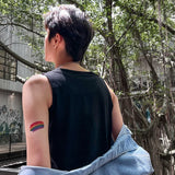 Close-up of a non-binary person with a short fade haircut, wearing a sleeveless black tee and denim jacket draped off the shoulders, revealing a rainbow Pride tattoo on their arm—an unapologetic symbol of LGBTQ+ identity and personal liberation.
