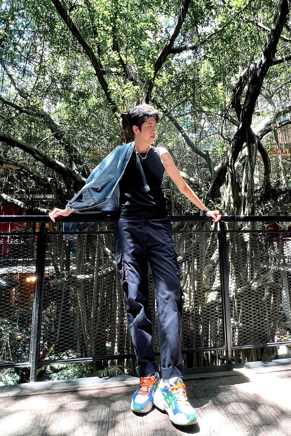 Androgynous individual in a black muscle tank and navy cargos leans confidently against a railing under a canopy of banyan trees, exuding genderfluid style and pride. TOMSCOUT's minimalist fit and bold stance echo queer freedom and urban nature vibes.