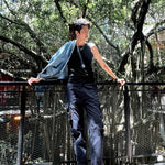 Androgynous individual in a black muscle tank and navy cargos leans confidently against a railing under a canopy of banyan trees, exuding genderfluid style and pride. TOMSCOUT's minimalist fit and bold stance echo queer freedom and urban nature vibes.