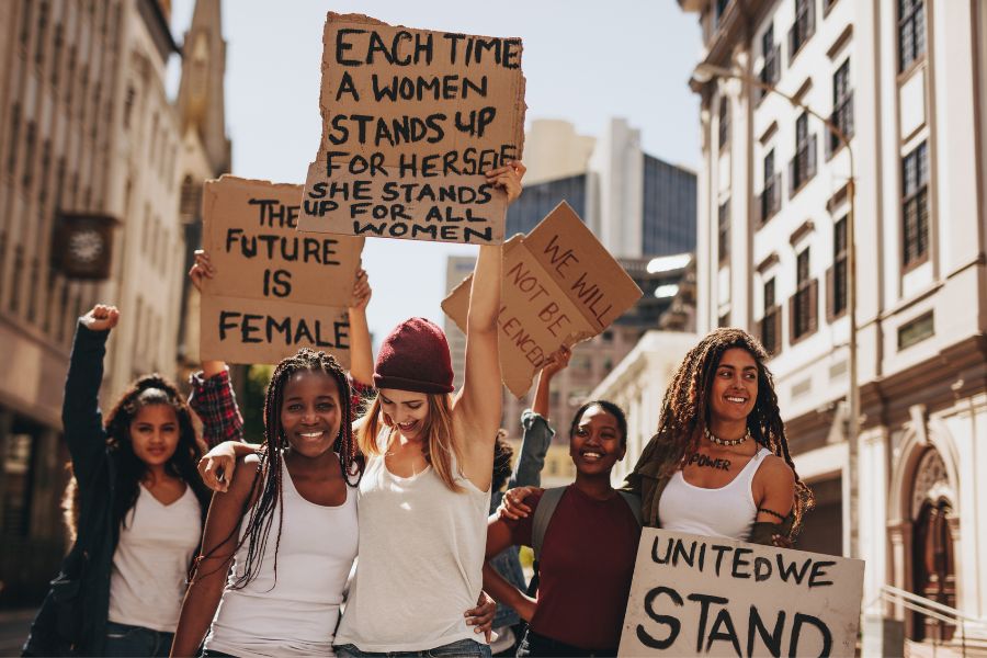 Joyful LGBTQ+ and feminist protestors holding signs like “United We Stand” and “The Future Is Female” in support of equality and trans visibility.