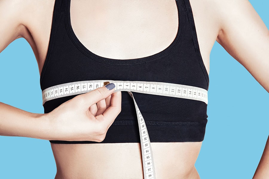 A woman in a black sports bra demonstrates how to measure her top bust with a measuring tape against a blue backdrop. TOMSCOUT Chest Binder Size Guide