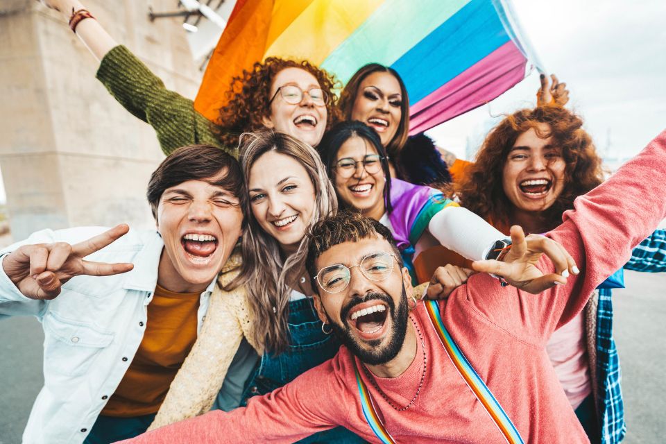 A diverse group of young people celebrating LGBT Pride Day, holding a pride flag together. Their joyful expressions and united stance reflect the spirit of inclusion and community, highlighting the vibrant energy of gay pride celebrations.