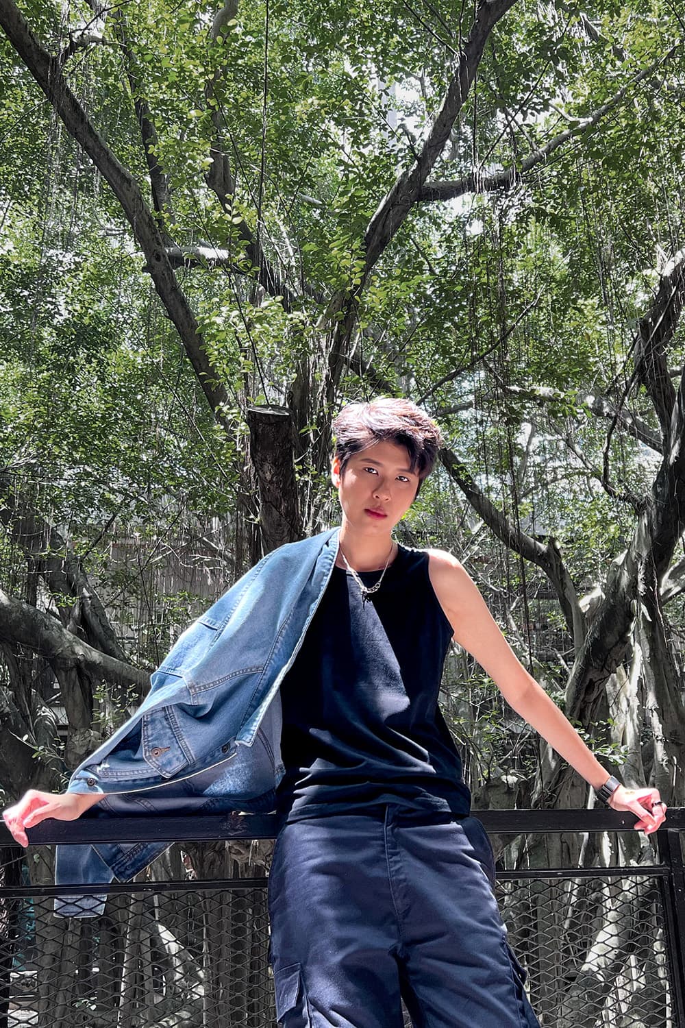 Lesbian model standing confidently under a lush canopy of trees, wearing a TOMSCOUT tank top chest binder and navy cargo pants with a denim jacket draped over one shoulder—radiating queer strength, tomboy elegance, and effortless urban cool in natural light.