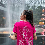 An Asian female with long hair is wearing a hot pink t-shirt that features a silk screen printing of 'love wins' on the back. She is standing in front of the Rain Vortex at Jewel Changi Airport. TOMSCOUT Flourish - LGBT Pride Kits (Singapore)