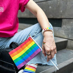An Asian female with long hair is wearing a hot pink t-shirt adorned with an embroidery design of a classic batik orchid flower on the front, along with various LGBT rainbow pride accessories, bracelet and holding a small pride flag. TOMSCOUT Flourish - LGBT Pride Kits (Singapore)