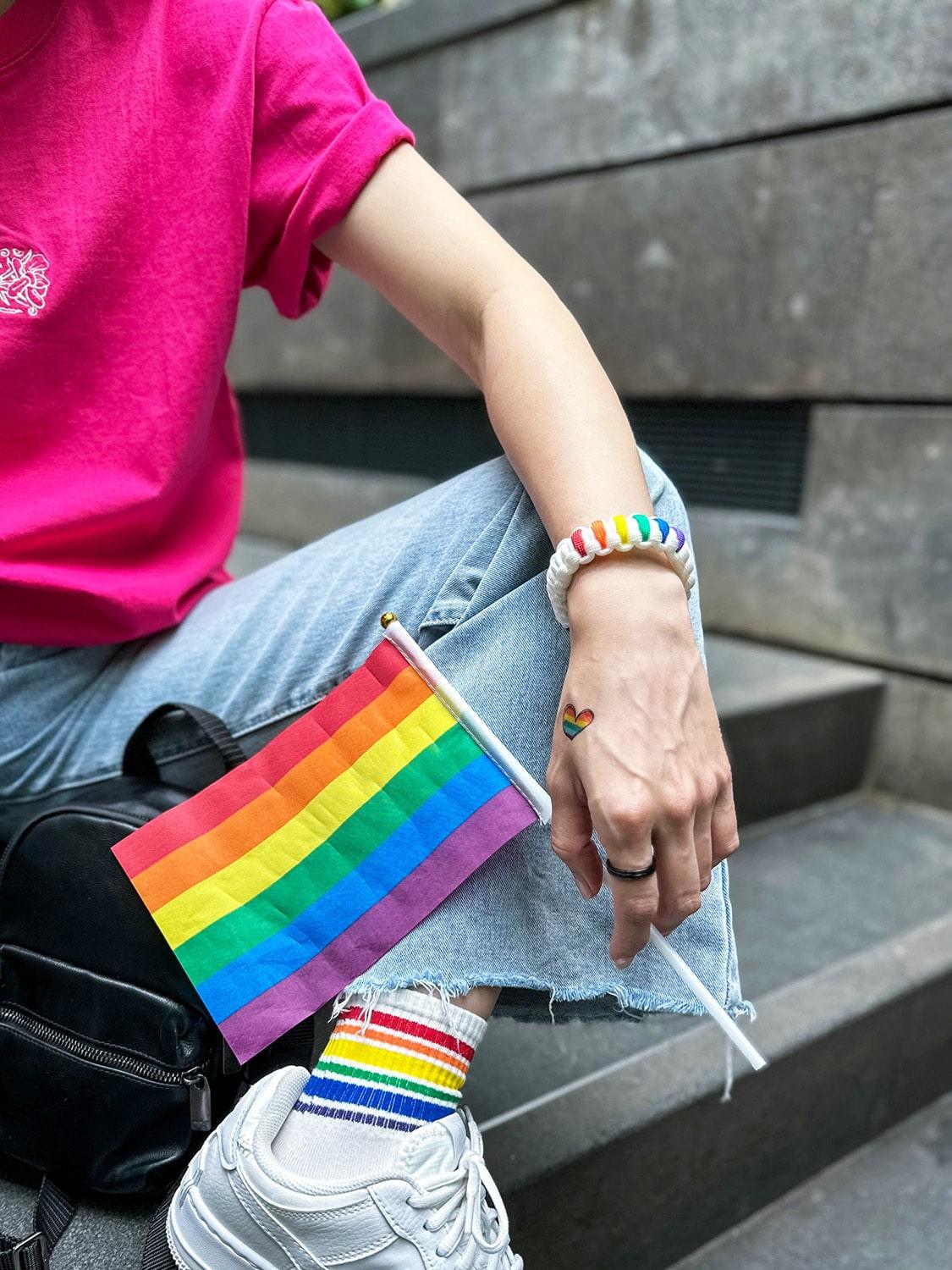 An Asian female with long hair is wearing a hot pink t-shirt adorned with an embroidery design of a classic batik orchid flower on the front, along with various LGBT rainbow pride accessories, bracelet and holding a small pride flag. TOMSCOUT Flourish - LGBT Pride Kits (Singapore)