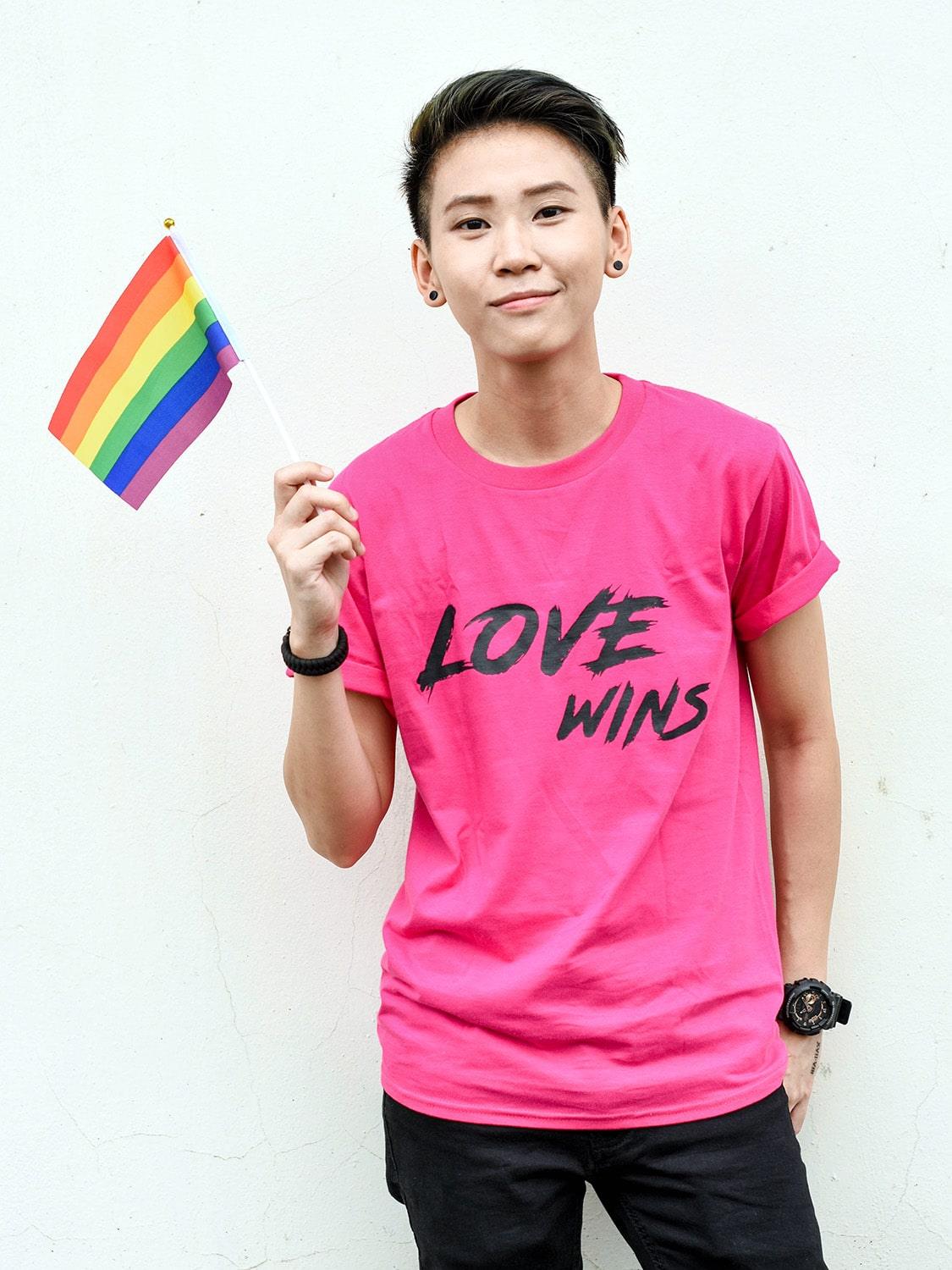 Captivating image of an Asian tomboy with a clean haircut, sporting a hot pink 'Love Wins' t-shirt and proudly holding a pride flag. Explore the TOMSCOUT Flourish - LGBT Pride Kits for a bold expression of pride in Singapore.