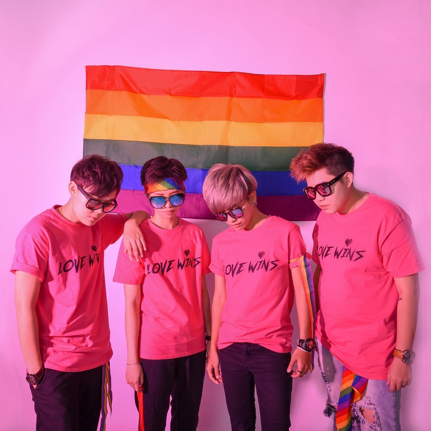Asian tomboys in hot pink 'Love Wins' t-shirts, posing against a large pride flag backdrop, embodying the spirit of LGBT pride. Discover TOMSCOUT Flourish - LGBT Pride Kits, a vibrant celebration of love and equality in Singapore.