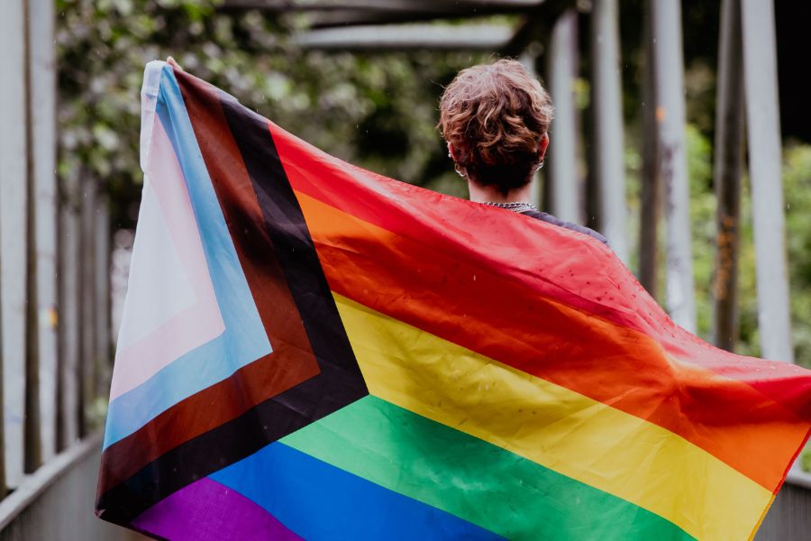 A transgender, non-binary person holding a progress pride flag outdoors. | TOMSCOUT chest binder body dysphoria, self love journey and self-acceptance journey
