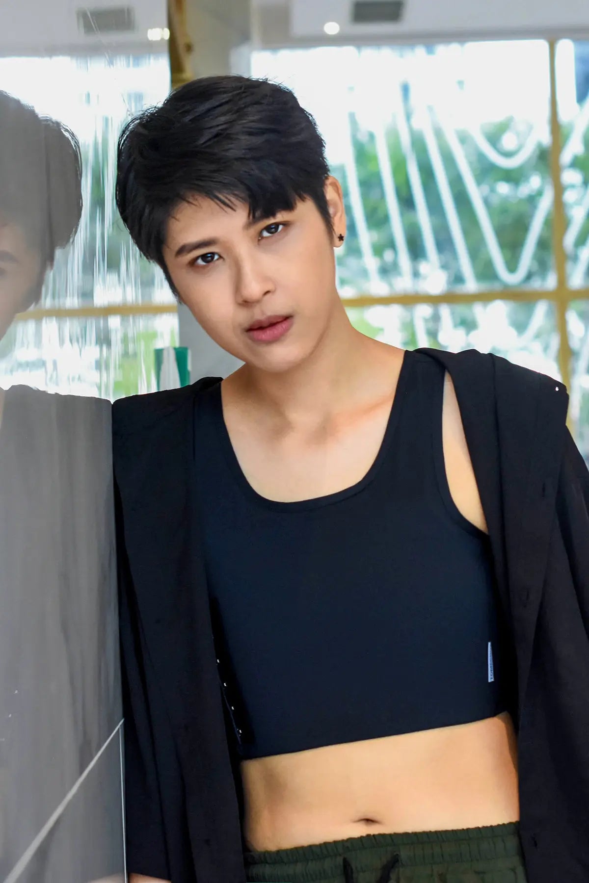 Close-up of a confident tomboy model wearing a sleek black TOMSCOUT chest binder, gazing assertively into the camera while leaning against a reflective surface in a bright, contemporary space—embodying gender euphoria, minimalist streetwear, and self-assured queer identity.