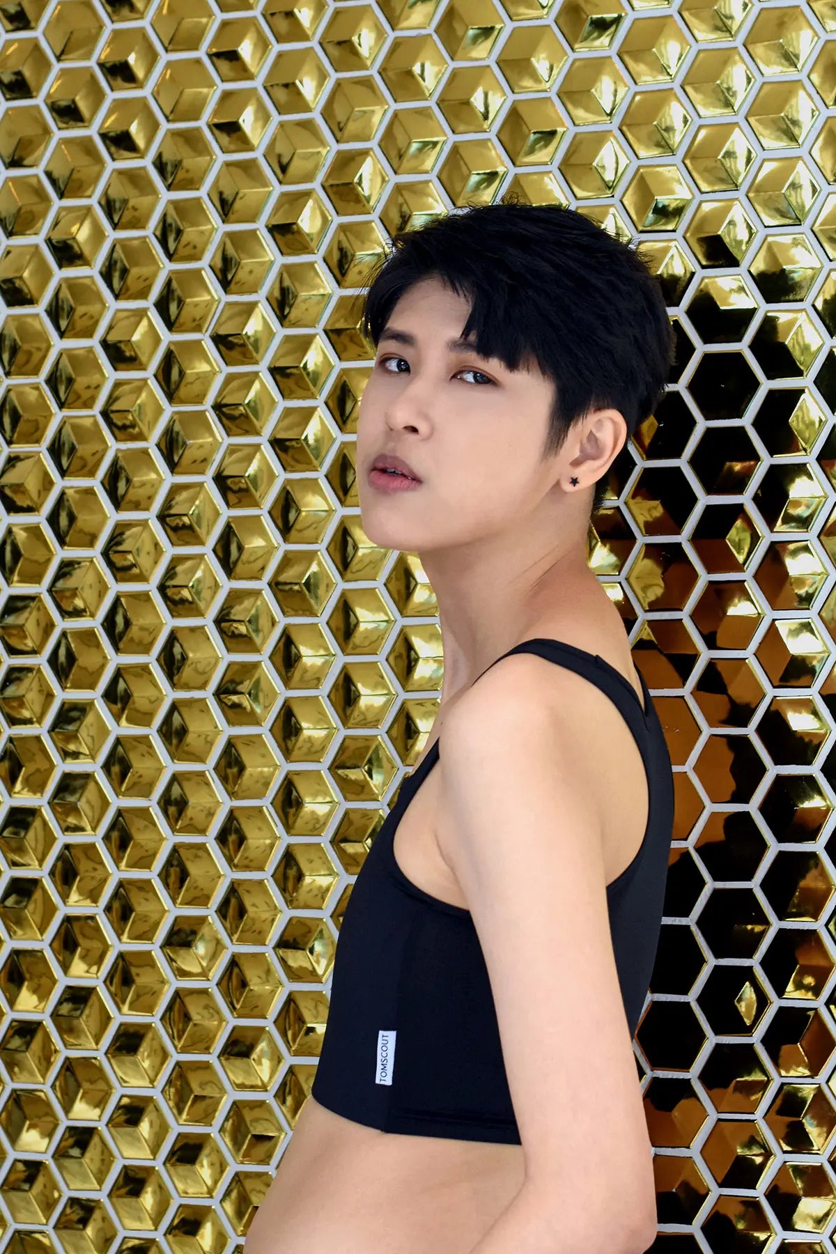 Androgynous model posing confidently in a black TOMSCOUT chest binder against a striking gold honeycomb wall, exuding sleek minimalism and genderfluid style with a subtle defiance in their gaze.