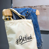 Close-up product image of a white 'Be Kind' embroidered canvas tote bag, adorned with an LGBTQ+ pride rainbow design, highlighting the details of the TOMSCOUT BE KIND Tote Bag.
