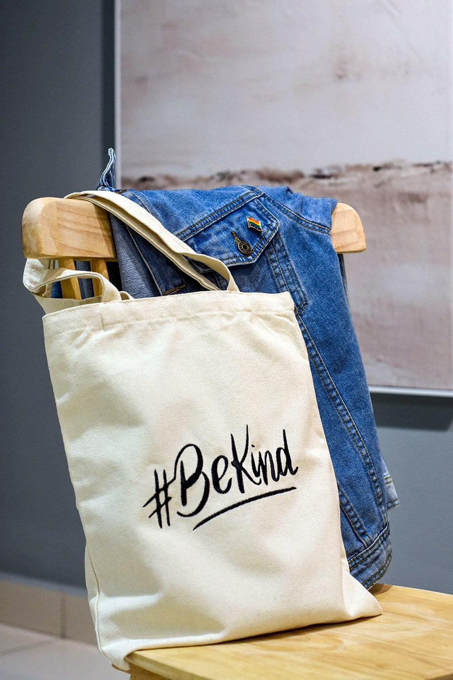 A white color embroidered Be Kind canvas tote bag with the LGBTQ+ pride black color design on the chair with a denim color jacket. TOMSCOUT BE KIND - Tote Bag