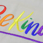 A white color embroidered Be Kind canvas tote bag with the LGBTQ+ pride rainbow color design. TOMSCOUT BE KIND - Tote Bag