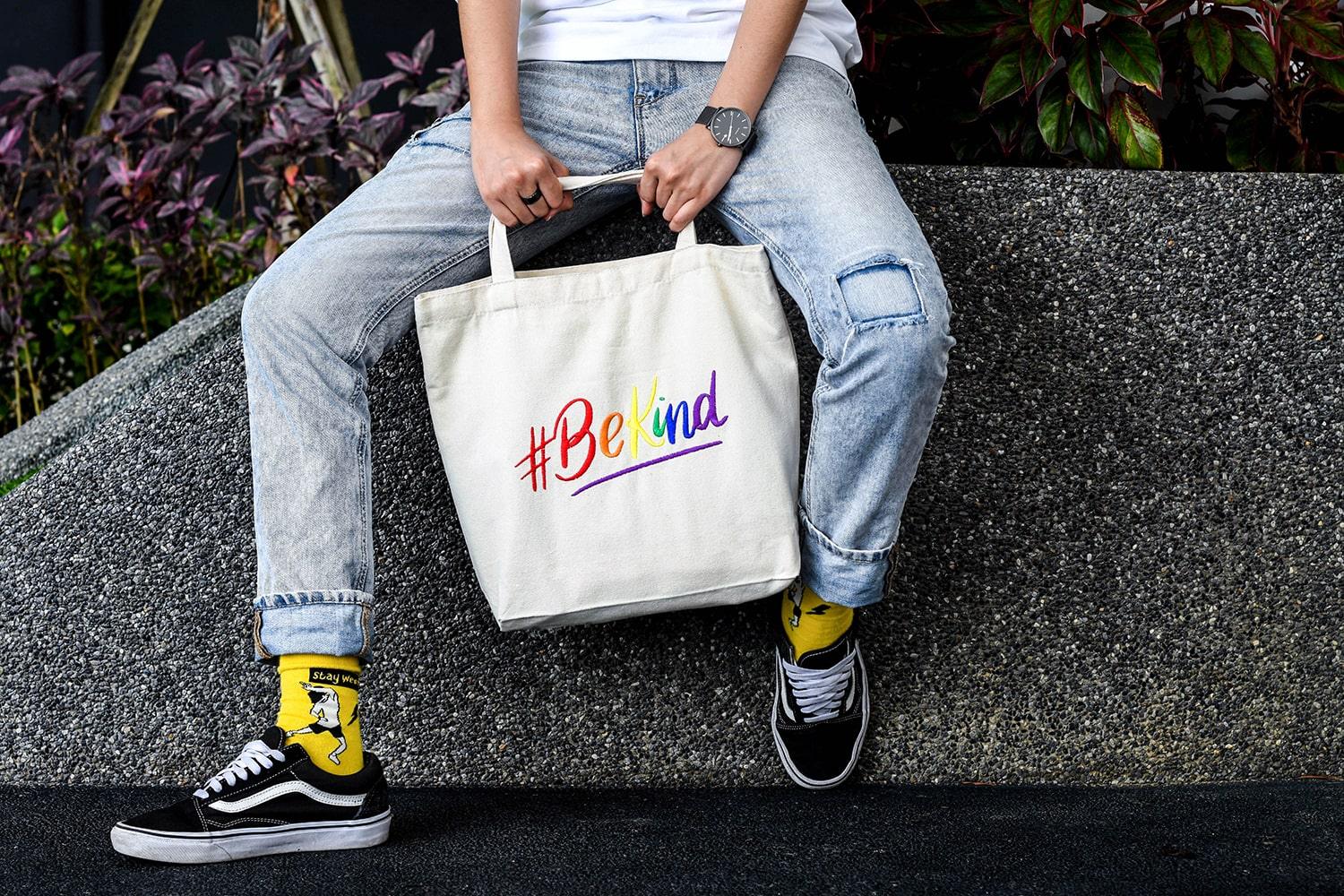 Close-up product image of a white 'Be Kind' embroidered canvas tote bag, adorned with an LGBTQ+ pride rainbow design, highlighting the details of the TOMSCOUT BE KIND Tote Bag.