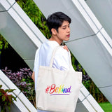 A non-binary androgynous tomboy, transgender man proudly holding a white color 'Be Kind' embroidered canvas tote bag with an LGBTQ+ pride black design, from the TOMSCOUT BE KIND Tote Bag collection.