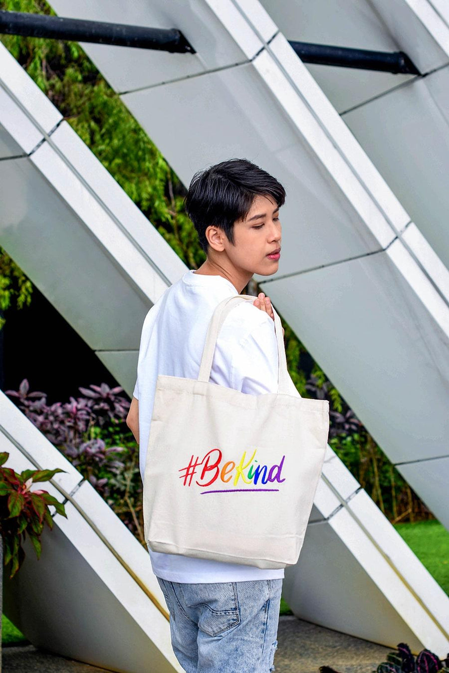 A non-binary androgynous tomboy, transgender man holding a white color embroidered Be Kind canvas tote bag with the LGBTQ+ pride rainbow color design. TOMSCOUT BE KIND - Tote Bag