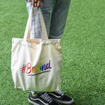 A non-binary androgynous tomboy, transgender man holding a white color embroidered Be Kind canvas tote bag with the LGBTQ+ pride rainbow color design. TOMSCOUT BE KIND - Tote Bag