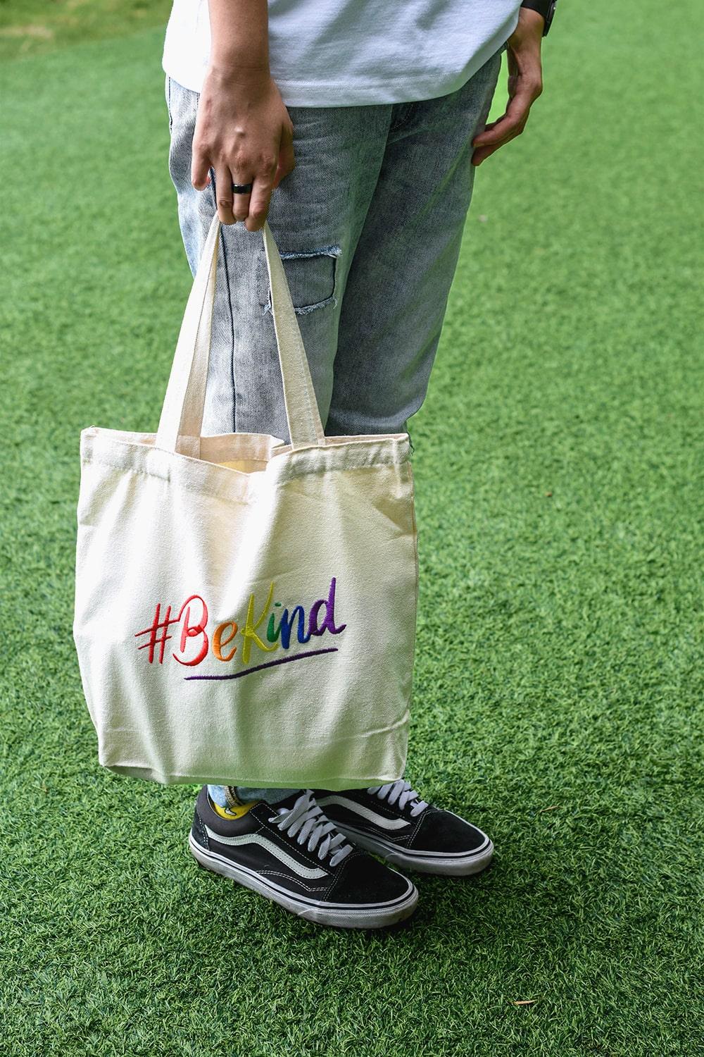 A non-binary androgynous tomboy, transgender man proudly holding a white color 'Be Kind' embroidered canvas tote bag with an LGBTQ+ pride black design, from the TOMSCOUT BE KIND Tote Bag collection.