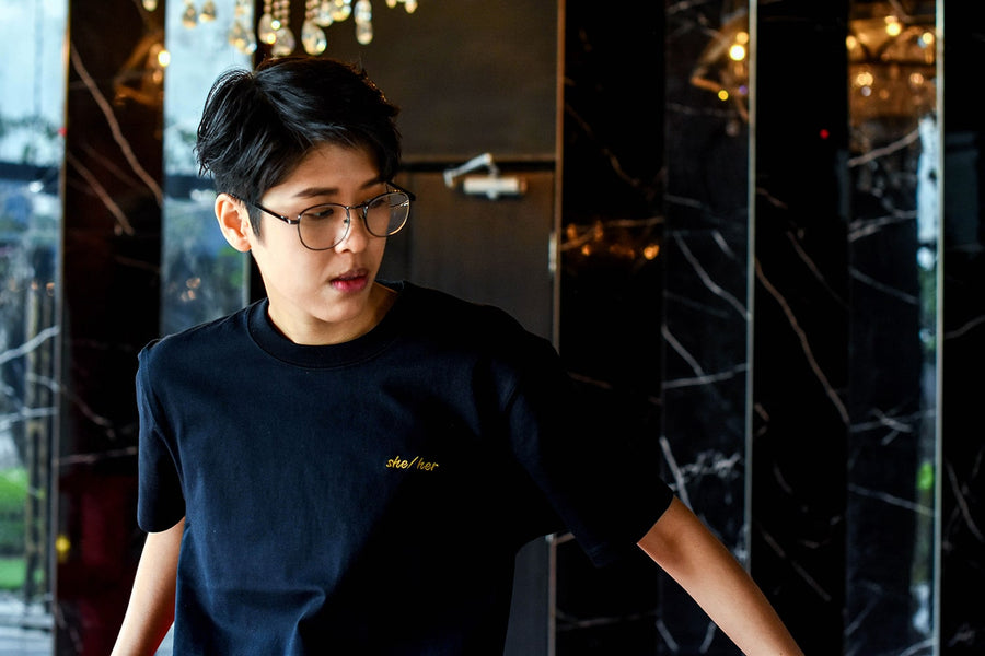A non-binary androgynous tomboy, transgender man wearing a black color t-shirts with embroidered gold color gender pronouns she/her on her left chest. TOMSCOUT Gender Pronouns she/her t-shirts - Customize Embroidered Clothing